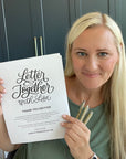 Woman holding a 'Letter Together with Lisa' book and pen set in front of a dark cabinet.